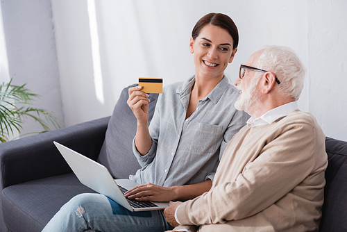 smiling woman holding credit card and laptop near aged father while sitting on sofa at home