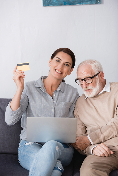 cheerful woman holding laptop and credit card while sitting near elderly father