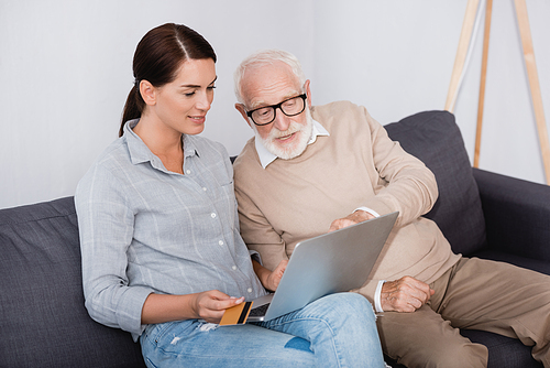 brunette woman holding credit card while using laptop together with elderly father