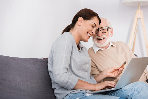 cheerful woman holding laptop and credit card near happy elderly father