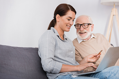 smiling woman using laptop and holding credit card near aged father at home