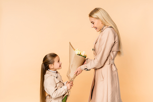 side view of cheerful kid giving flowers to happy mother on 8 march isolated on beige