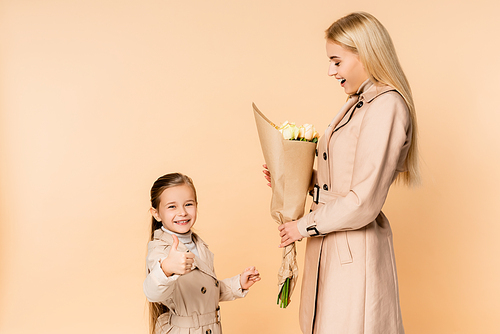cheerful kid showing thumb up near amazed mother holding flowers on 8 march isolated on beige