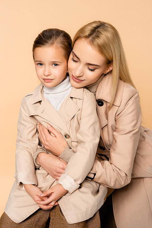 caring mother hugging daughter in trench coat isolated on beige