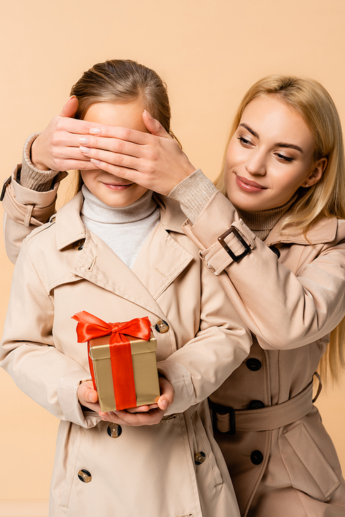 cheerful mother covering eyes of kid with gift box isolated on beige