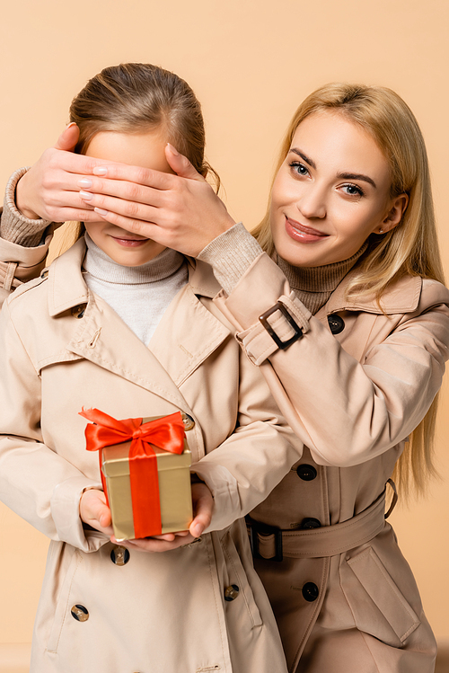 happy mother covering eyes of kid with gift box isolated on beige