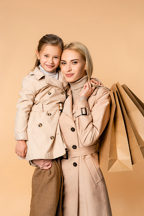 smiling mother holding paper bags and hugging daughter isolated on beige
