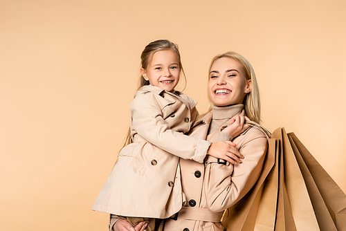 smiling mother holding paper bags and hugging cheerful daughter isolated on beige