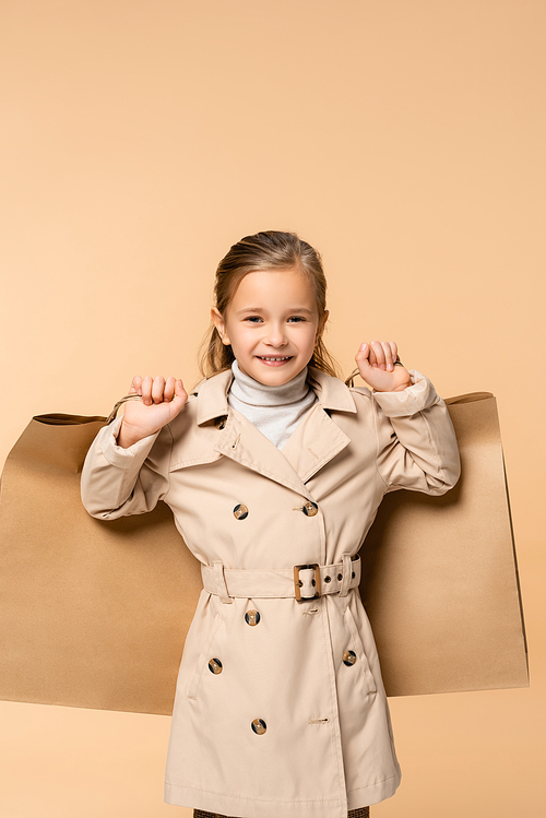 happy kid in trench coat holding paper bags isolated on beige