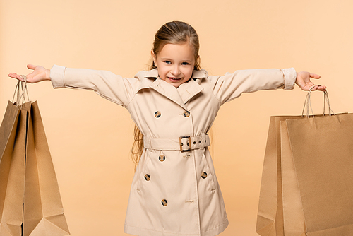 cheerful kid in trench coat holding paper bags isolated on beige