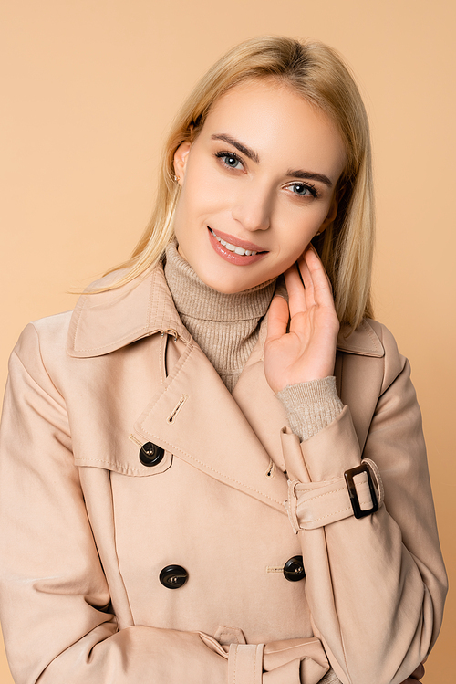 blonde woman in trench coat smiling and  isolated on beige