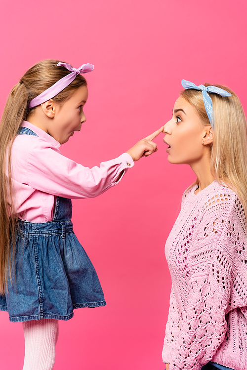 side view of surprised kid touching nose of mother isolated on pink