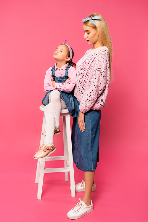full length of mother looking at offended kid sitting on chair on pink