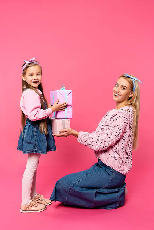happy mother smiling near daughter holding presents on pink