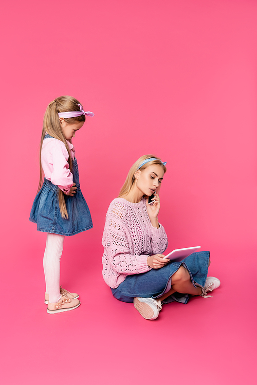 offended kid looking at mother talking on smartphone and using digital tablet on pink