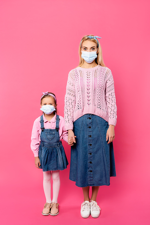 full length of mother and daughter in medical masks standing and holding hands on pink