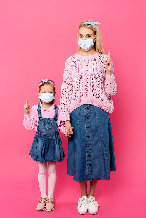 full length of mother and daughter in medical masks pointing with fingers and holding hands while standing on pink