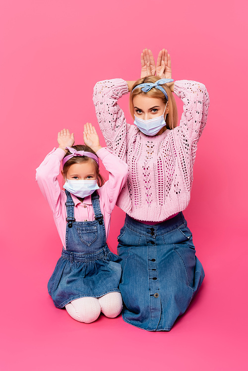 mother and daughter in medical masks making bunny ears with hands on pink