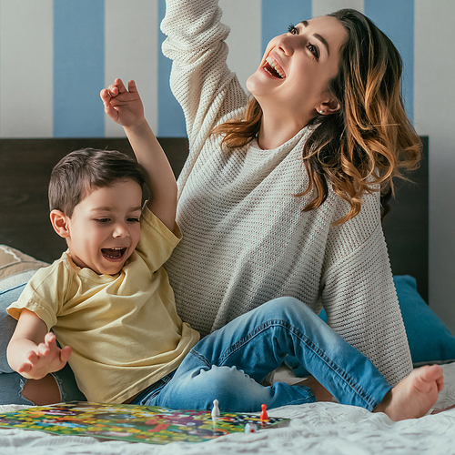 happy, excited mother and son showing winner gestures while playing board game on bed