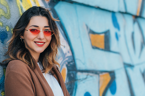 stylish woman in sunglasses smiling and standing near wall with graffiti