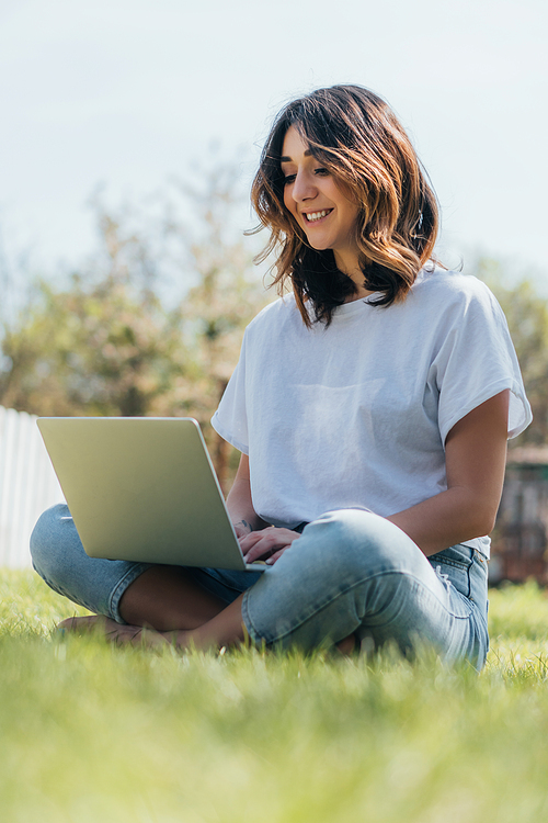 selective focus of happy freelancer using laptop and sitting on green grass