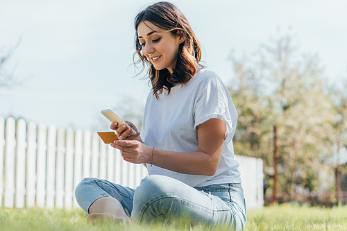 selective focus of happy woman sitting on grass, holding credit card and smartphone while online shopping