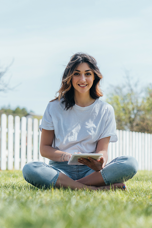 selective focus of happy woman sitting on grass and holding digital tablet