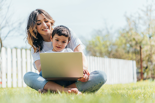 selective focus of happy mother and son using laptop while sitting on grass