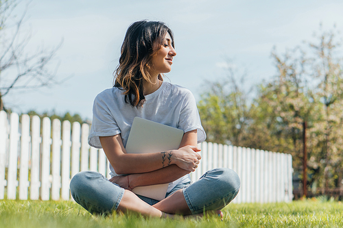 selective focus of cheerful woman sitting on grass and holding laptop