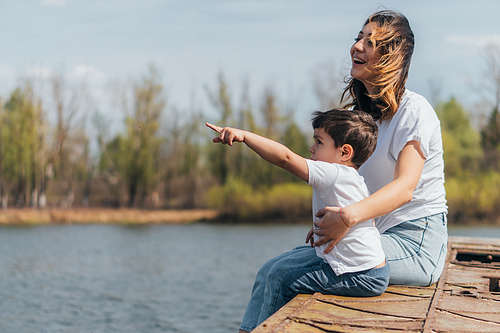 cute kid pointing with finger while sitting near happy mother and lake