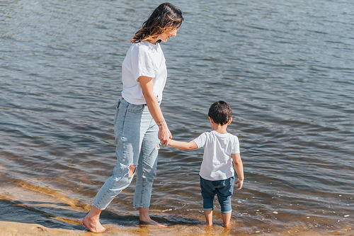 happy mother holding hands with cute son and standing in lake