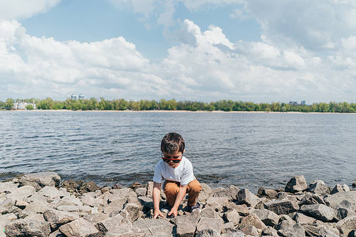 stylish boy in sunglasses touching rock while sitting near lake