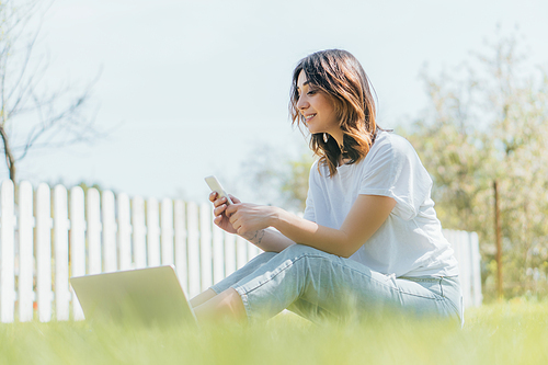 selective focus of happy woman using smartphone near laptop while sitting on grass