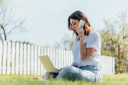 selective focus of happy woman talking on smartphone and using laptop while sitting on grass
