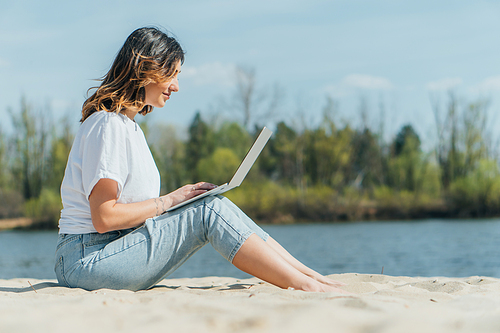 side view of happy freelancer sitting on sand and using laptop