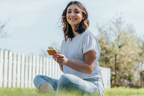 selective focus of cheerful woman sitting on grass, holding credit card and smartphone while online shopping