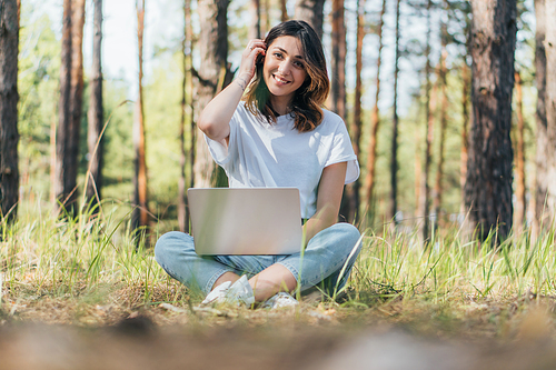 selective focus of happy freelancer sitting with laptop in woods