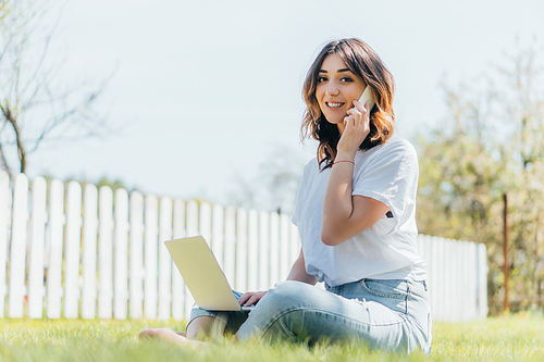 selective focus of attractive freelancer talking on smartphone near laptop and sitting on grass