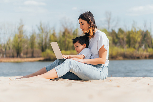 selective focus of mother and son sitting on sand and using laptop