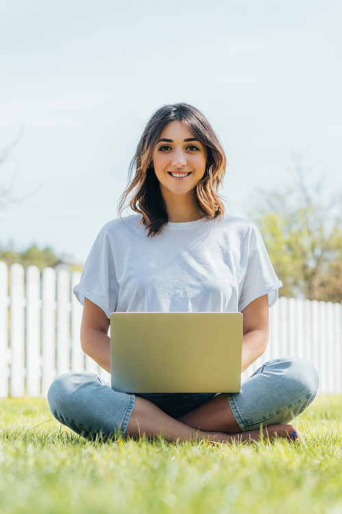 selective focus of smiling freelancer using laptop and sitting on grass