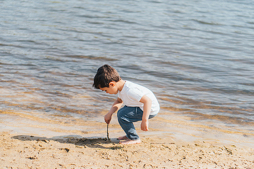 barefoot boy holding stick near wet sand and pond