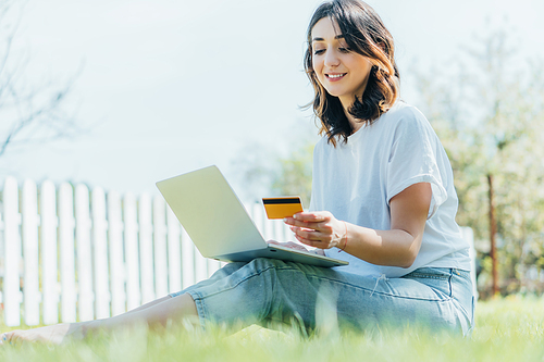 selective focus of happy woman looking at credit card near laptop and sitting on grass