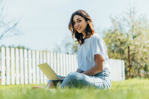 selective focus of smiling woman using laptop and sitting on grass