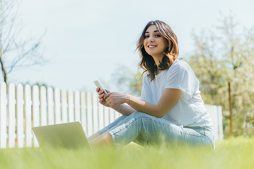 selective focus of cheerful woman holding smartphone near laptop and sitting on grass
