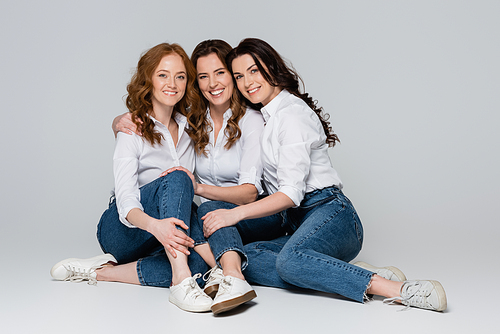 Adult women in jeans embracing and smiling at camera on grey background