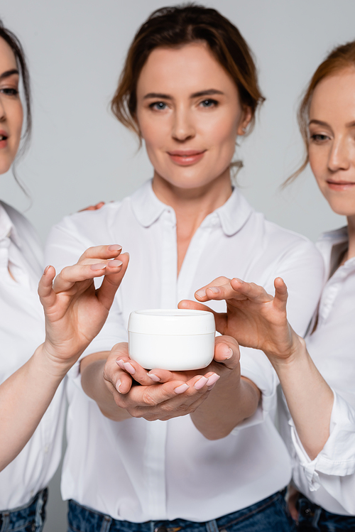 Jar with cosmetic cream in hand of woman near friends on blurred background isolated on grey