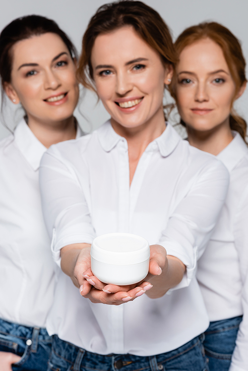 Jar with cosmetic cream in hands of smiling women on blurred background isolated on grey