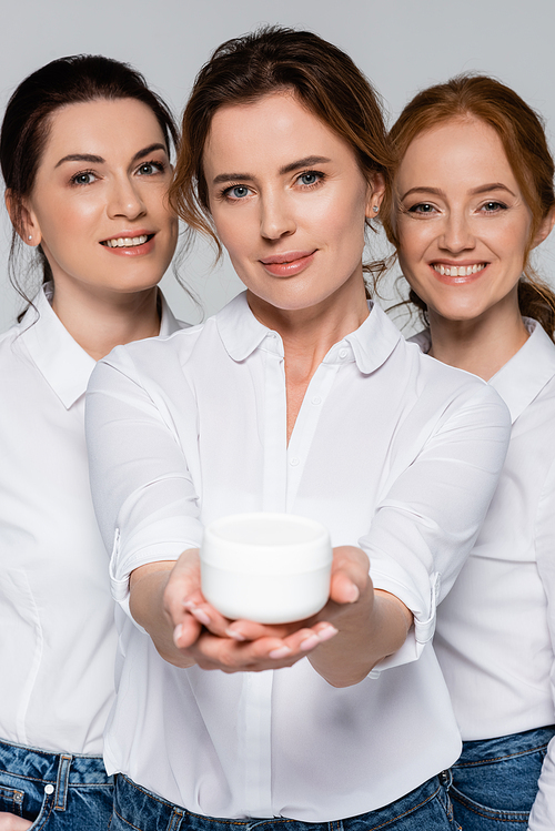 Smiling women  near friend with jar of cosmetic cream on blurred foreground isolated on grey