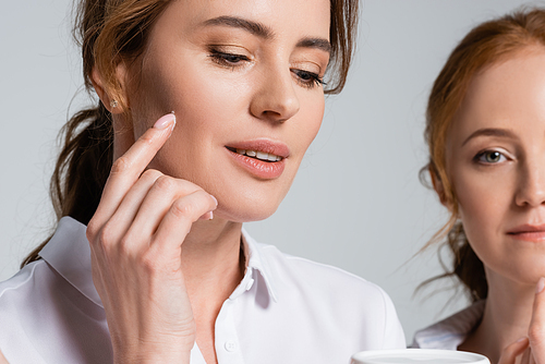 Woman applying face cream near friend on blurred background isolated on grey