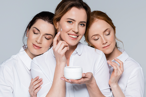 Smiling woman applying face cream near friends with closed eyes isolated on grey